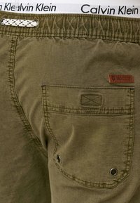 Olive green shorts with a textured finish, featuring a back pocket with stitching details, metal eyelets, and a white waistband branded "Calvin Klein."