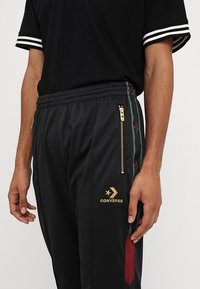 Mies, który ma na sobie czarne spodnie dresowe z złotym logo Converse, zielonymi i czerwonymi bocznymi paskami oraz kieszeniami na zamek, a do tego czarną koszulkę w paski na rękawach.