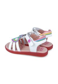 Sandalias infantiles blancas con múltiples correas, un tacón con acentos en rosa y arcoíris, y una suela roja texturizada. Diseño colorido destacado en la parte superior.