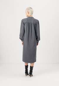 Lang, grå denimkjole med puffermer og en samlet bærestykke. Brukt med svarte hælhøye støvler. Glatt tekstur og knelangs snitt.