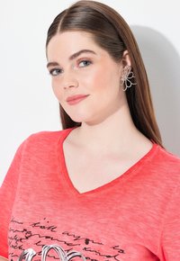 Camiseta roja de cuello en V con una superficie texturizada y diseño impreso en negro; grandes pendientes florales plateados con bucles intrincados y acentos.