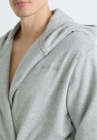 Pilkas gobtuvas apsiaustas, pagamintas iš minkštos medžiagos, su šalikėlio apykakle ir iš dalies atvirais priekiais. Diskretiškas „Calvin Klein“ logotipas išsiuvinėtas ant krūtinės.