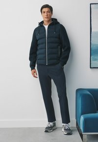 Doudoune marine avec capuche et zip, associée à un pantalon sombre et des baskets grises. Texture lisse, design ajusté, subtils accents en métal.
