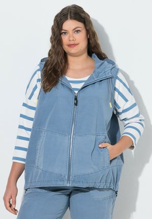 Mujer con cabello largo y ondulado que lleva una chaqueta azul sin mangas con cremallera sobre una camisa de manga larga blanca y azul a rayas, de pie con una mano en el bolsillo.