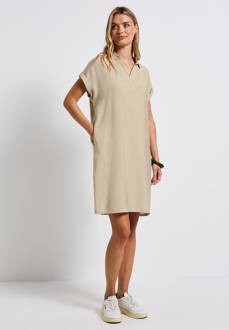 Beige poloklänning med korta rullade ärmar och sidofickor, gjord av lättviktsmaterial, når ovanför knäet, stylad med vita sneakers.