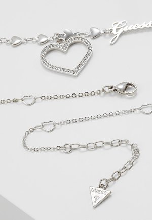 Pulsera de charms de plata con un colgante en forma de corazón con cristales, eslabones en forma de corazón y un charm triangular con el logo "Guess" sobre un fondo blanco.