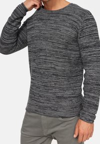 Homme portant un pull tricoté texturé gris et noir à manches longues et un pantalon gris, une main près de son cou.