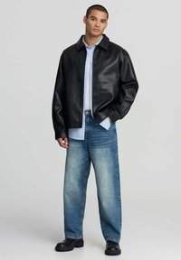 Giovane uomo con i capelli corti, che indossa una giacca di pelle nera, una camicia azzurra chiara, jeans blu sbiaditi e stivali neri, in piedi con una mano in tasca.