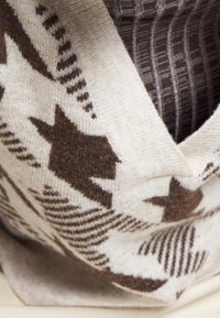 Zbliżenie na beżowy i brązowy dziergany sweter z wzorami gwiazd i geometrycznymi, noszony na ciemnej, prążkowanej koszuli.