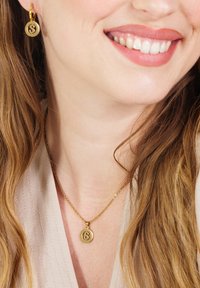 Gouden ketting met een cirkelvormige hanger met een gestileerde "S," vergezeld van bijpassende oorbellen met een hoop. Gladde textuur, gepolijste afwerking.