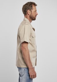Camicetta beige a maniche corte con colletto button-down, due taschini sul petto e orlo dritto, abbinata a jeans in denim azzurri chiari.