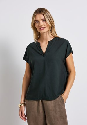 Haut court à manches courtes de couleur vert foncé avec un décolleté en V et des détails de couture subtils au col. Tissu lisse ; coupe décontractée, porté avec un pantalon beige.