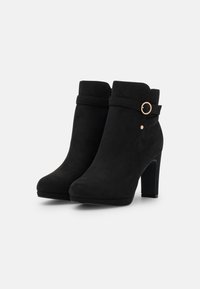 Schwarze Wildleder-Stiefeletten mit runder Zehenpartie, hohem Blockabsatz und dekorativem Riemen mit einem goldenen Ringsdetail nahe der Oberseite.