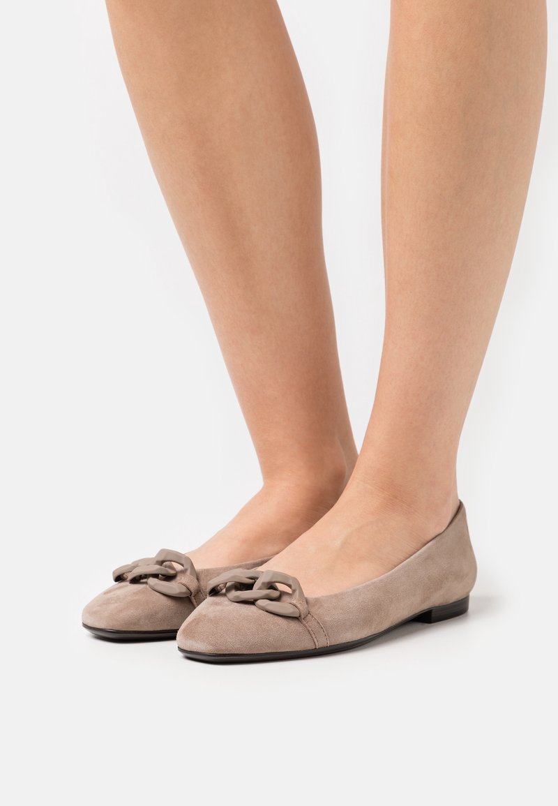 Pieds portant des ballerines en daim taupe avec un grand détail de chaîne assorti sur l'orteil arrondi, sur un fond blanc uni.