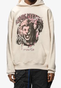 Sudadera con capucha en color blanco roto, con un diseño gráfico de un león rugiente acompañado de motivos de rayos y el texto "¿Artista o Asesino?" en la parte inferior.