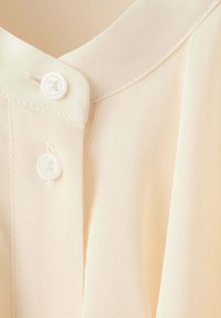 Gros plan d'un col de chemise couleur crème avec deux boutons blancs et une couture visible le long du bord.