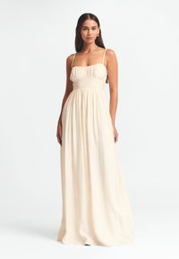 Langes, cremefarbenes Kleid mit enganliegendem Oberteil, gerafftem Taillenbund und schmalen Trägern. Hergestellt aus einem leichten, strukturierten Material.