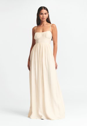 Langes, cremefarbenes Kleid mit enganliegendem Oberteil, gerafftem Taillenbund und schmalen Trägern. Hergestellt aus einem leichten, strukturierten Material.