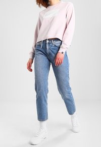 Světle růžová zkrácená mikina s bílou obdélníkovou přední nášivkou, kombinovaná se světle modrými kalhotami s vysokým pasem a bílými teniskami.