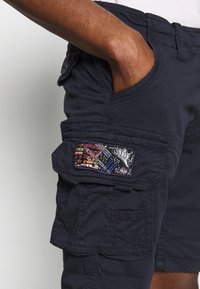Shorts cargo bleu marine en tissu texturé, poche latérale avec broderie colorée représentant des paysages urbains et un thème spatial, et coupe à taille standard.