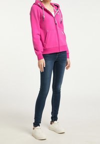 Fuchsiová mikina na zip s kapucí na šňůrku, dvěma předními kapsami a kontrastními detaily šňůrky, spárovaná s úzkými modrými džíny a bílými teniskami.