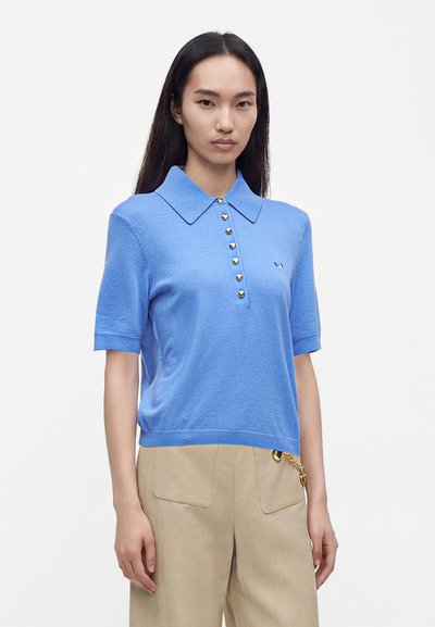 Femme aux longs cheveux foncés portant un polo tricoté bleu clair à manches courtes avec des boutons dorés et un pantalon beige avec un détail en chaîne dorée.
