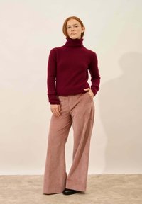 Młoda kobieta z rudymi włosami, ubrana w bordowy sweter z golfem i różowe spodnie z materiału sztruksowego o szerokich nogawkach, stojąca na jednolitym tle.