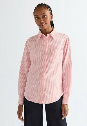 Młoda kobieta z zaplecionymi włosami, ubrana w różowo-białą koszulę w pionowe paski i ciemne spodnie, delikatnie się uśmiechająca na jednolitym tle.