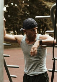 Un débardeur gris sans manches, un short de sport noir et une casquette noire. La personne interagit avec des équipements de gym métalliques dans un cadre extérieur.
