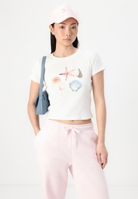 White, cropped t-shirt featuring colorful shell and starfish designs. Paired with pink joggers and a light pink cap. Blue shoulder bag.