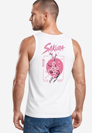 Mann trägt weißes Tanktop mit pinker Kirschblüte und Text "SAKURA" auf dem Rücken, kombiniert mit blauen Jeans, seitlich stehend.