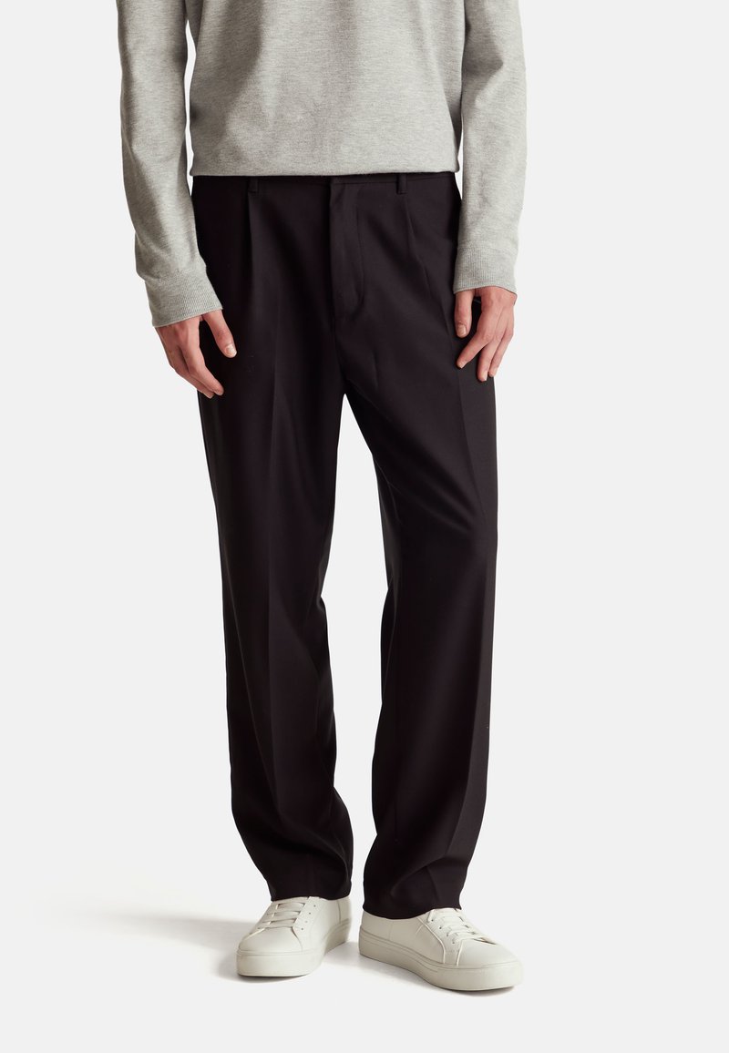 Černé kalhoty rovného střihu s předními sklady, vyrobené z hladké tkaniny. Nošeny s svetrem světle šedé barvy a bílými teniskami.