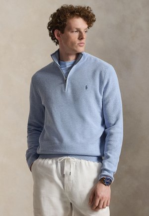 Unge mand med krøllet hår iført en lyseblå kvartlynlås trøje, hvide bukser med snøre, og et armbåndsur med blå urskive, kigger til siden.