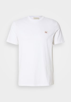 T-shirt blanc à manches courtes et col rond avec un petit patch logo Calvin Klein rouge et beige sur le côté gauche de la poitrine.