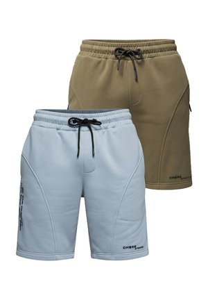 Homme debout portant un t-shirt jaune clair, un short bleu clair, des chaussettes blanches et des baskets blanches sur un fond gris uni.