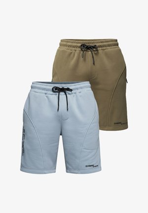 Mann steht und trägt ein hellgelbes T-Shirt, hellblaue Shorts, weiße Socken und weiße Sneakers vor einem einfarbigen grauen Hintergrund.