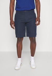 Marineblå shorts lavet af vævet stof, med knaplukning, forlommer, flad front og lige pasform, parret med hvide sneakers.
