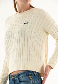 Suéter de punto color crema con un patrón de trenzas, cuello y dobladillo acanalados, que presenta un pequeño logo en el pecho. Combinado con jeans azules.