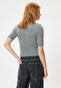 Grijze gebreide trui met korte mouwen en kabelpatroon, parelaccenten op de schouders, gecombineerd met hoge taille zwarte jeans, gezien van achteren.