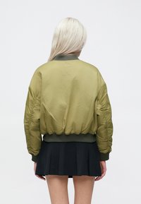 Olivengrønn bomberjakke med ribbestrikket kant og mansjetter; polstret design; lett skinnende tekstur; kombinert med et svart plissert skjørt.