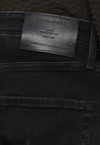 Crne traperice od trapera s kožnom oznakom na kojoj je utisnut tekst. Oznaka prikazuje marku "JACK & JONES" i dodatne informacije.