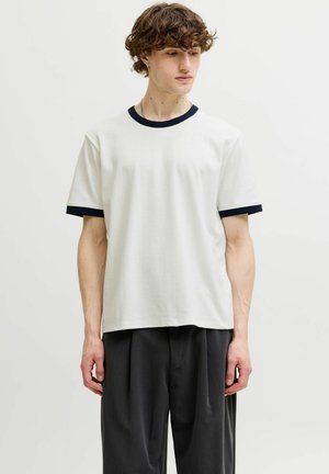 Ung mand med krøllet hår iført en hvid T-shirt med sort krave og ærmekanter, kombineret med løse sorte bukser, stående mod en hvid baggrund.