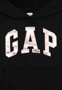 Sweatshirt à capuche noire avec un grand logo "GAP" en rose gold métallique sur le devant. Texture lisse avec une poche frontale.