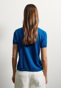 Maglietta blu a maniche corte in maglia con colletto rotondo e texture liscia, abbinata a pantaloni chiari. Design semplice, senza motivi o dettagli.