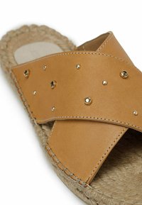 Brune lædersandal med krydset stropper, prydet med metalliske nitter og en juteindpakket sål. Glat tekstur med syede detaljer.