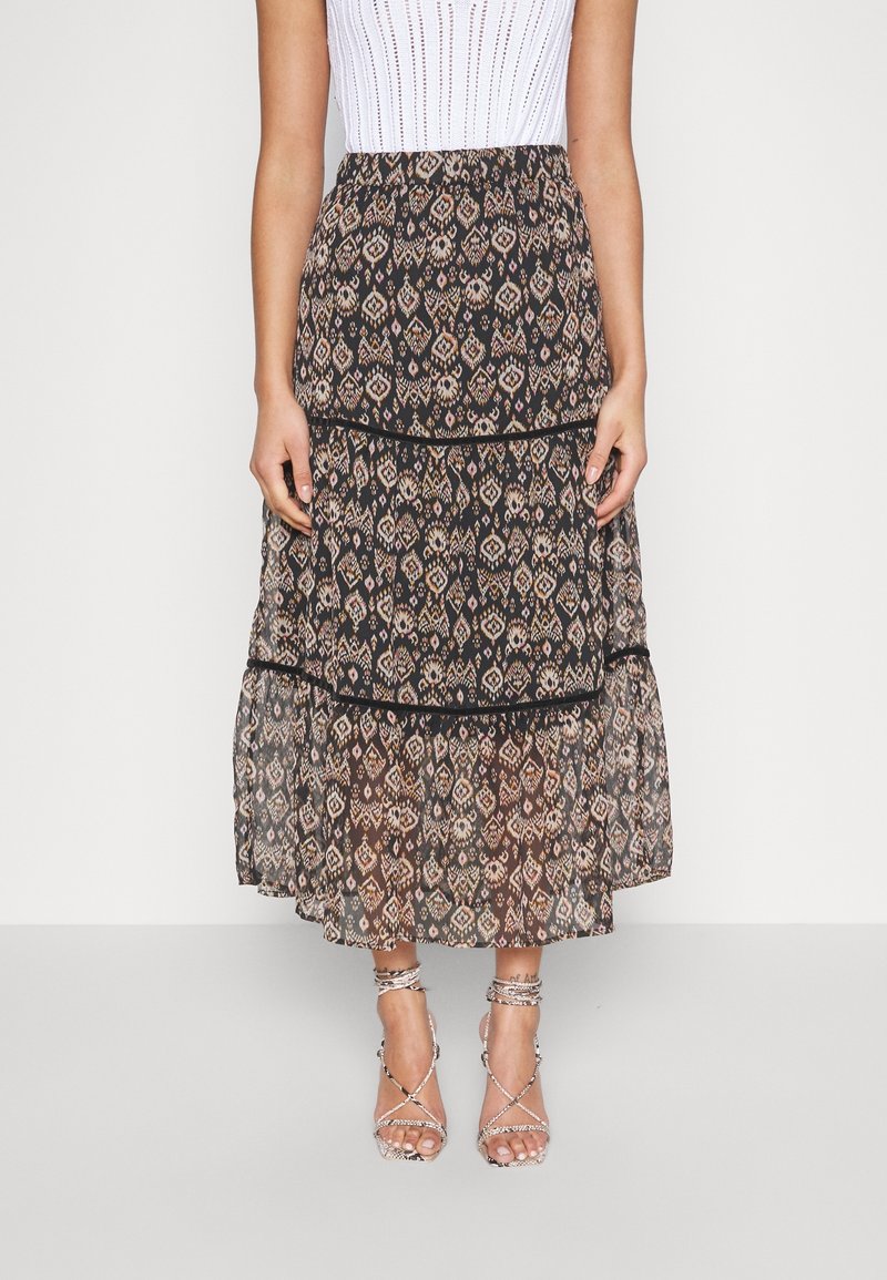 Jupe maxi noire à motifs avec ourlet à volants superposés, présentant des dessins floraux et géométriques complexes, associée à des sandales argentées à brides.