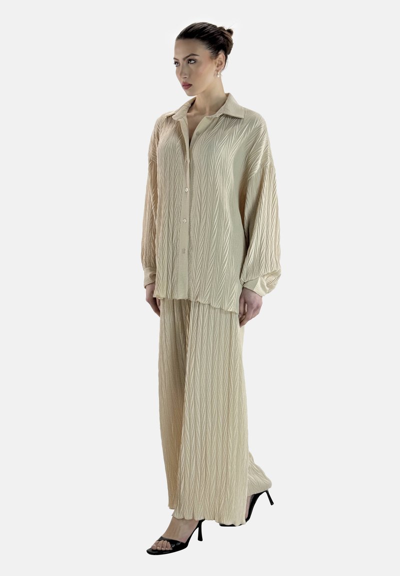 Ensemble deux pièces texturé beige comprenant une chemise longue à boutons avec col et un pantalon à jambes larges, complété par des sandales à talons noires.