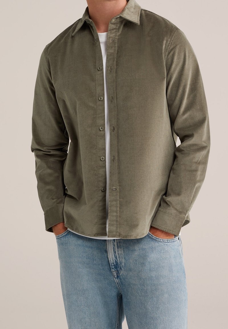 Homme portant une chemise en velours côtelé vert olive déboutonnée par-dessus un t-shirt blanc avec un jean bleu clair, les mains dans les poches.