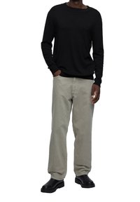 Homme portant une chemise noire à manches longues, un pantalon ample gris clair et des bottes noires, debout une main dans la poche sur fond blanc.