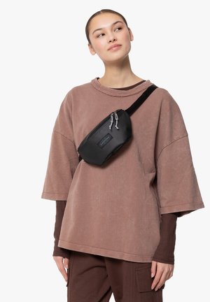 Zwarte nylon heuptas met twee ritssluitingen, gedragen over het lichaam boven een oversized, stoffige rozen sweatshirt.